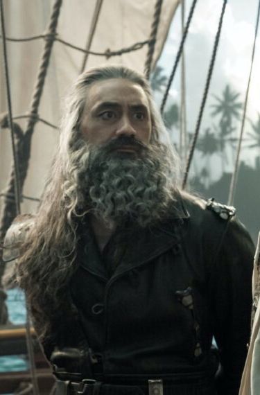 Edward Teaxh in season 1 episode 9 of Our Flag Means Death, wearing his Blackbeard leathers with his hands restrained behind his back, standing on the main deck of the Revenge and looking at Stede out of frame with a concerned expression