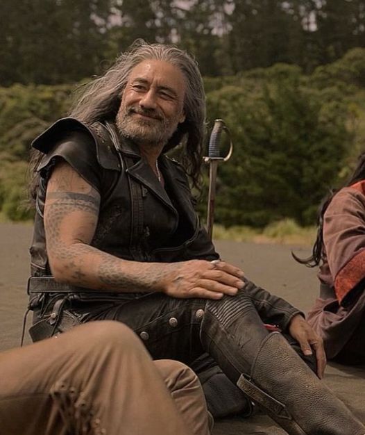 Edward Teach in season 2 episode 8 of Our Flag Means Death, wearing his Blackbeard leathers, sitting on a beach in the Republic of Pirates and looking at Stede out of frame beside him with a fond, proud smile