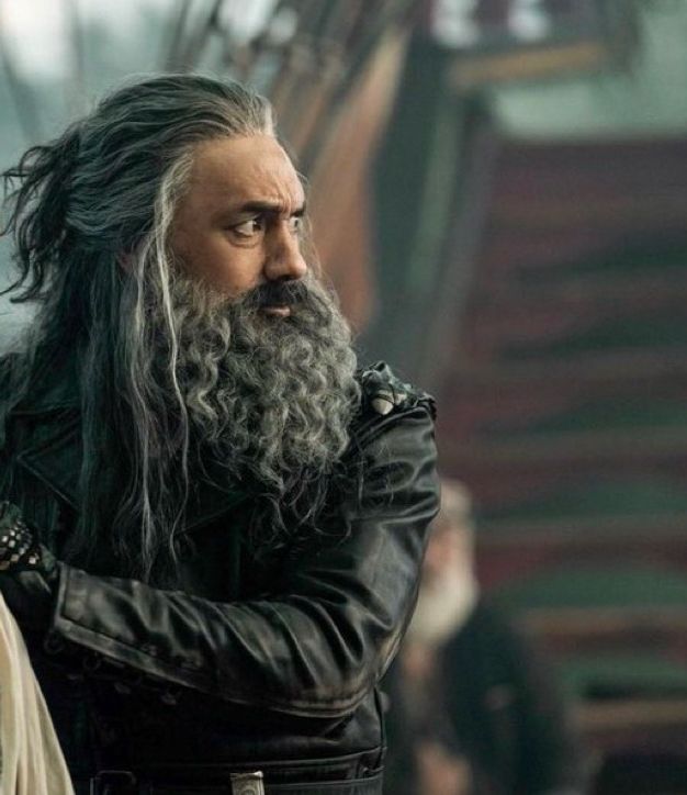 Edward Teach in season 1 episode 9 of Our Flag Means Death, wearing his Blackbeard leathers, standing on the main deck of the Revenge protectively in front of Stede out of frame with a worried expression