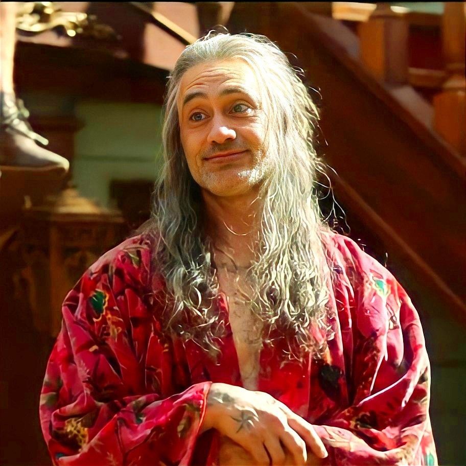 Edward Teach in season 1 episode 10 of Our Flag Means Death, wearing a fuchsia tropical bird-print robe, standing on the main deck of the Revenge and looking at the crew out of frame with a soft smile 