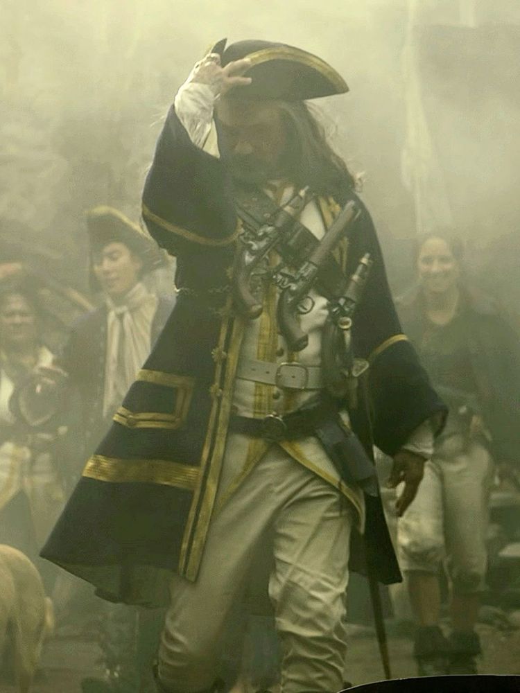 Edward Teach in season 2 episode 8 of Our Flag Means Death, wearing an English Navy uniform with his head tilted down as he puts on a hat and walks through fog in the Republic of Pirates