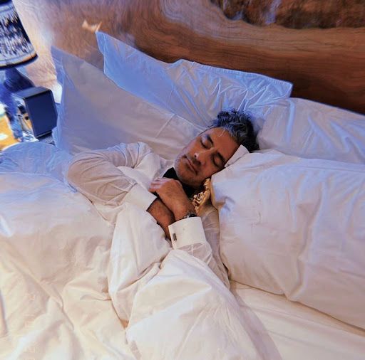 Taika Waititi wearing a white button-up shirt and a black bow tie, laying in a bed and snuggling his Oscar with his eyes closed