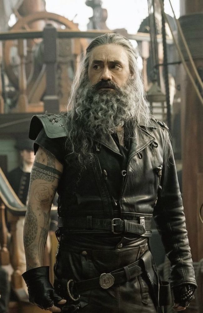Edward Teach in season 1 episode 9 of Our Flag Means Death, wearing his Blackbeard leathers and black leather fingerless gloves, standing on the main deck of the Revenge and looking over at Stede out of frame with a concerned expression