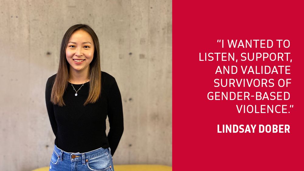 “I wanted to listen, support, and validate survivors of gender-based violence.” – Lindsay Dober
Photo: a smiling Lindsay Dober; she is wearing jeans, a black sweater and necklace, standing in front of a concrete wall.  