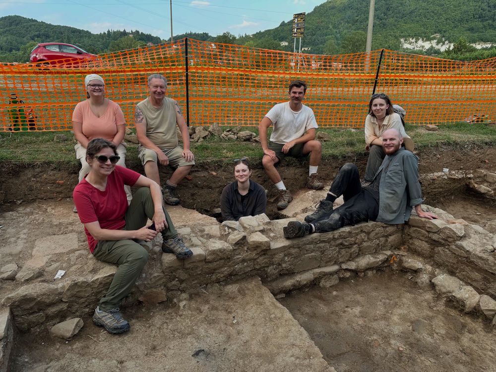 A group of six BSU students with Director Katie Huntley sitting on the walls in a trench. 