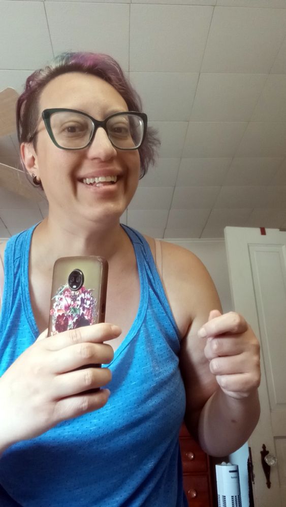 Me now, a Latine person in green cat-eye glasses and a blue tank top, holding a cell phone with art on the back of it of a skull covered in flowers. I'm in a similar position to the original photo. 
