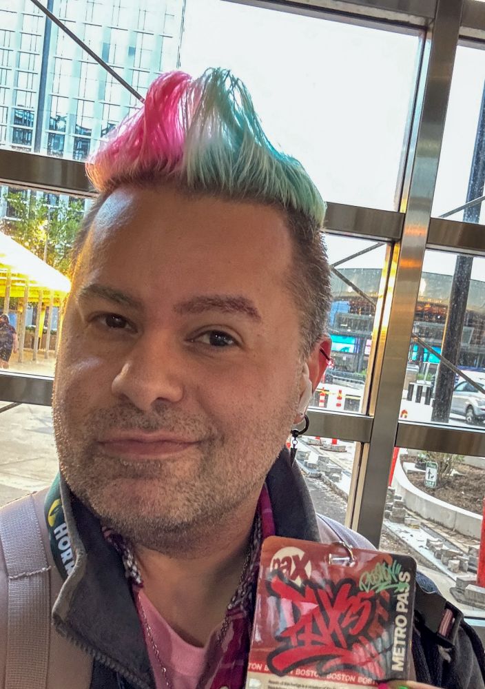 Ony, holding up their freshly-acquired PAX East badge, with the Boston Convention & Exhibition Centre looming ominously in the background.
