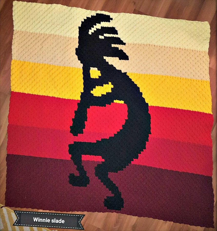 A handmade crochet afghan featuring a bold Kokopelli silhouette in black against a gradient background of warm sunset colors, including shades of yellow, red, and brown.