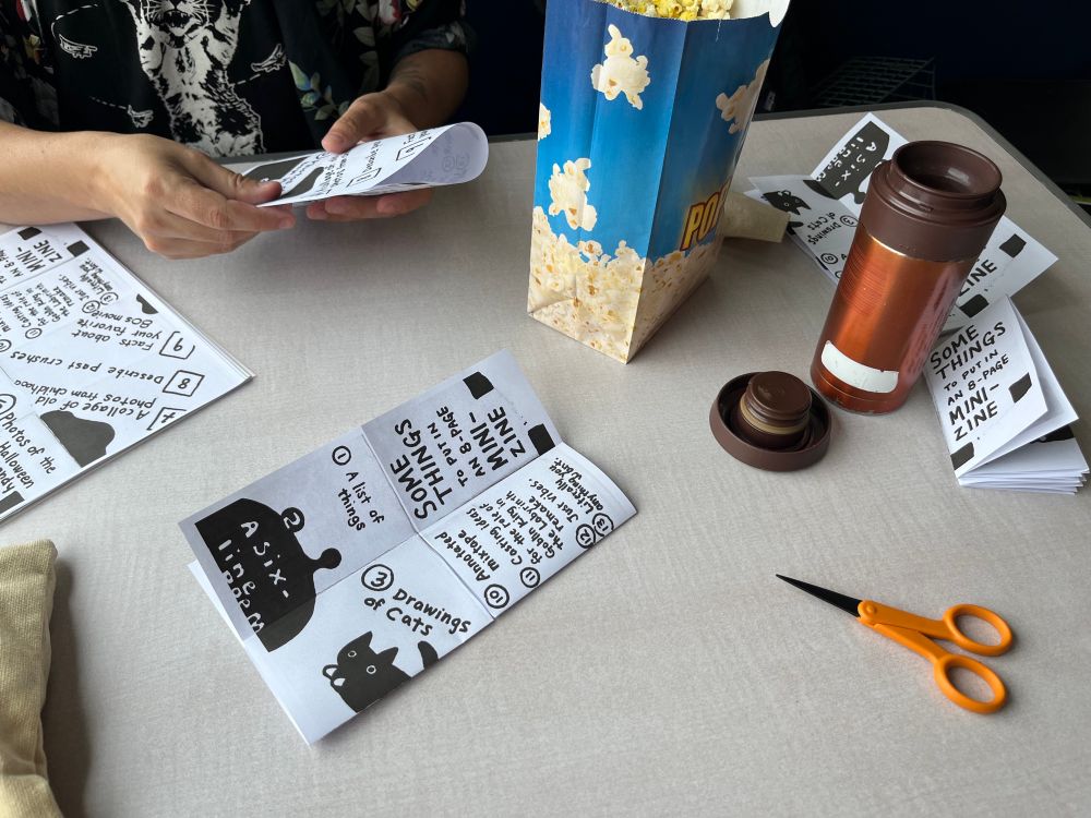 Photo of my partner and me folding mini zines on the ferry (and a bag of popcorn and mug of coffee)