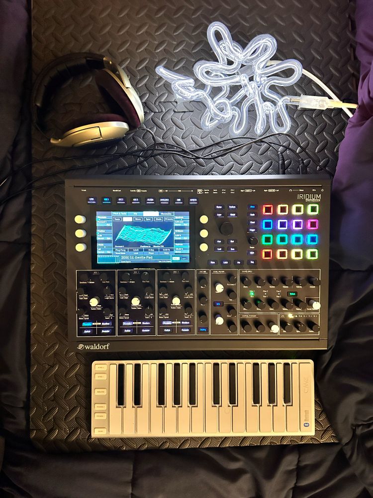 Apparently a now fully functioning Waldorf Iridium Desktop synthesizer with headphones attached as well as a CME Xkey Air 25 keyboard (via USB) along with a neon light sculpture next to it.