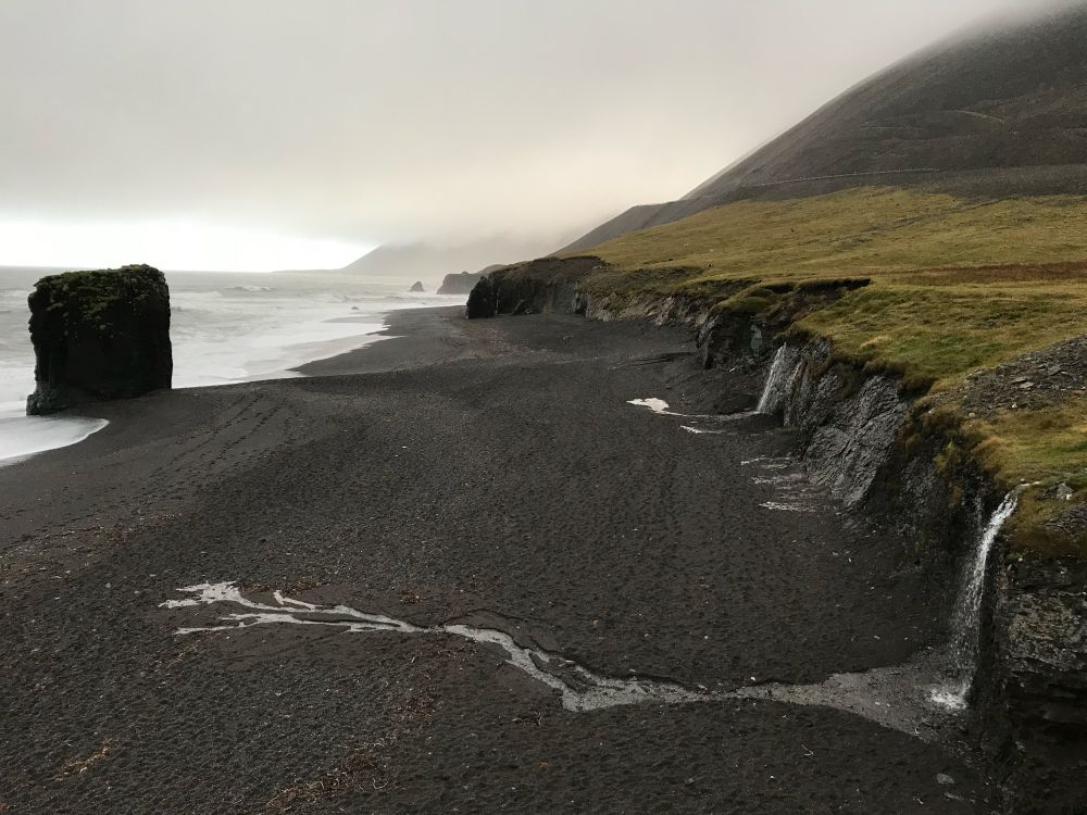 Black sand beach on the east coast of Iceland, late November, in the grey-clouded cold. There’s a large monolithic rock tower just off the beach in the water on the left, and on the right there are numerous small runoff waterfalls flowing down from a small cliff separating the land from the beach. The largest of these is in the foreground, and the runoff has almost but not quite made it to the ocean. The beach, cliff, and ocean go off miles into the distance eventually fading into the distant fog. There’s not a human in sight, just mossy highland, short cliff, narrow black sand beach, monolith, ocean, and fog. For endless miles.