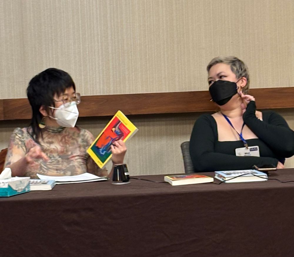 Two Asian American scholars at a conference table. Jina Kim on the left in a print top holding a brightly colored book while Khuc in a dark top looks on. Both scholars are masked, and looking at each other.