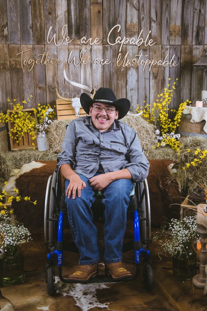 A young man in western wear and cowboy boots smiles enthusiastically. He's in a wheelchair and surrounded by barn dance decor such as hay and cow hides. The wall displays the phrase: We are Capable, Together We are Unstoppable. 