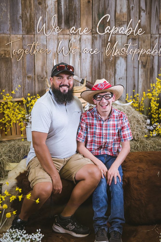 A young man in western wear with dark rimmed glasses and a cowboy hat smiles enthusiastically as he sits next to his father. They are surrounded by barn dance decor such as hay and cow hides. The wall displays the phrase: We are Capable, Together We are Unstoppable. 
