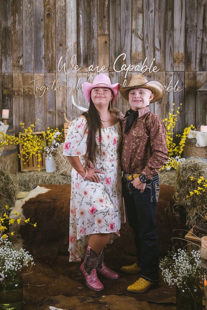 A young couple in western wear and cowboy boots pose together with joyful smiles. They are surrounded by barn dance decor such as hay and cow hides. The wall displays the phrase: We are Capable, Together We are Unstoppable. 