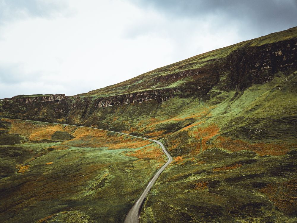 A narrow road winds through the center and then off to the left of green and rust-colored grasses and shrubs. The sky is cloudy and it hangs above a diagonal of cliffs.