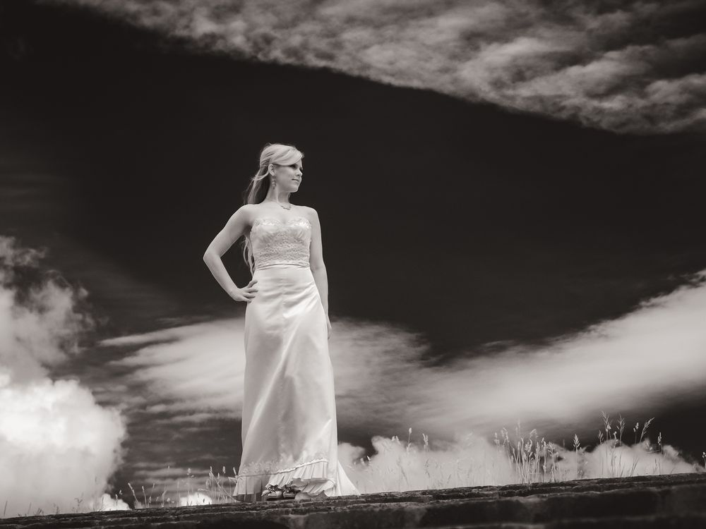 A blonde bride with long hair looking over her shoulder with sweeping clouds behind her. Taken a a low angle from below.