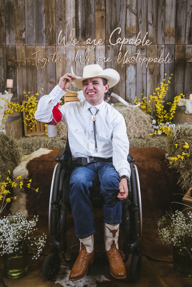 A young man in western wear and cowboy boots smiles enthusiastically and tips his cowboy hat. He's in a wheelchair and surrounded by barn dance decor such as hay and cow hides. The wall displays the phrase: We are Capable, Together We are Unstoppable. 