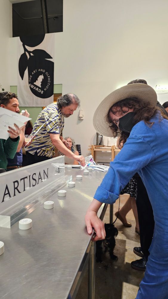 A cutie with light skin, a wide brimmed hat, curly brown hair and denim blue outfit leaning on a steel table in front of a row of perfume samples.
