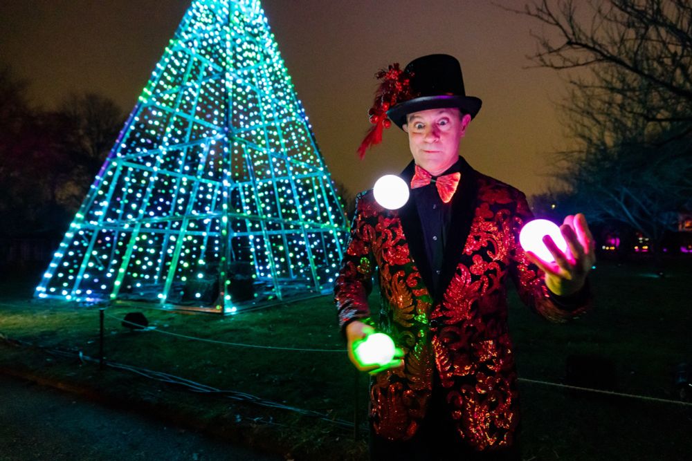 A performer in dapper suited costume juggles lighted balls outdoors in front of a light sculpture in the shape of an abstract tree.