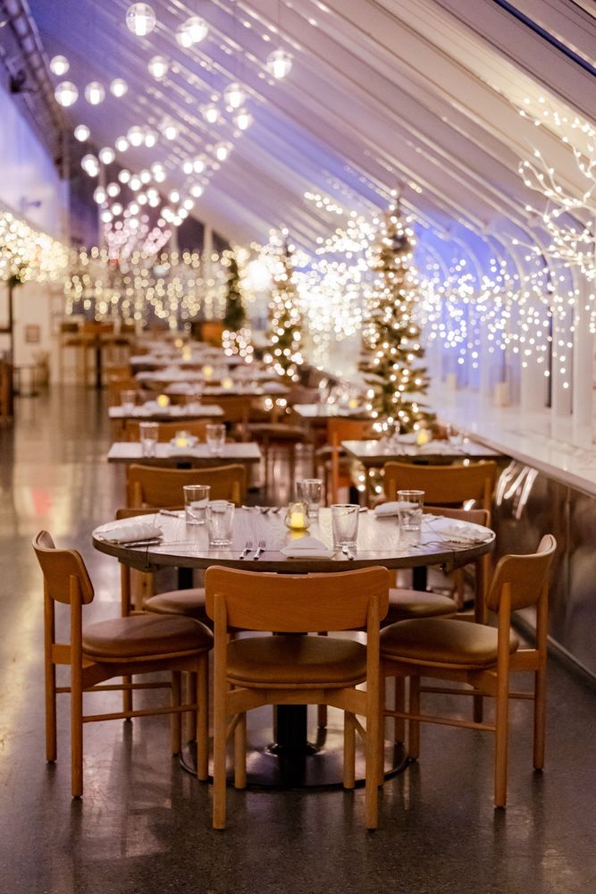 A restaurant inside a glassy curved ceiling with many circular tables and many many lights and wintery accents.