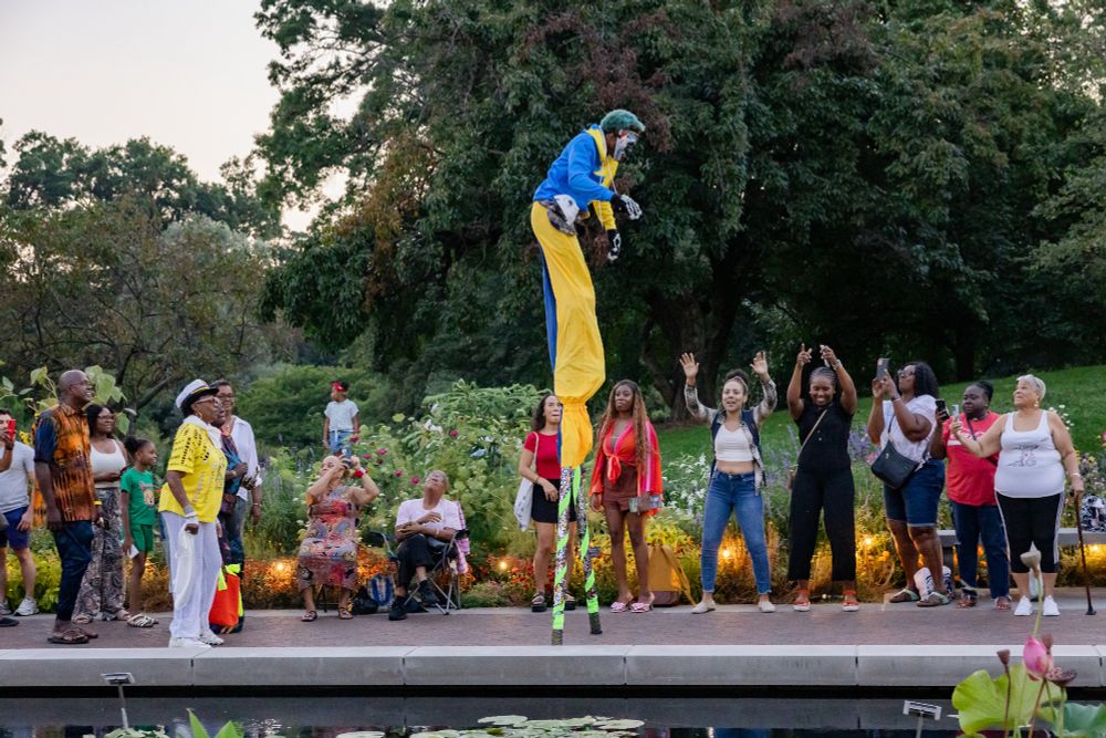 A person on stilts wearing a clown outfit and makeup walks amidst a group of excited people who stand on a path inbetween a planting of summer plants illuminated by small lights and a water feature.