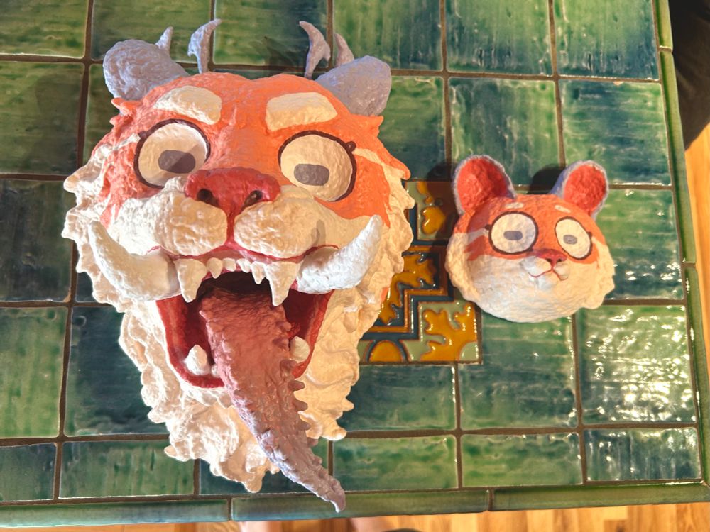 two side-by-side papier maché masks of Percy from Cosmoknights. he is a purple and coral-colored tiger with two forms- a little cute guy and a big scary guy
