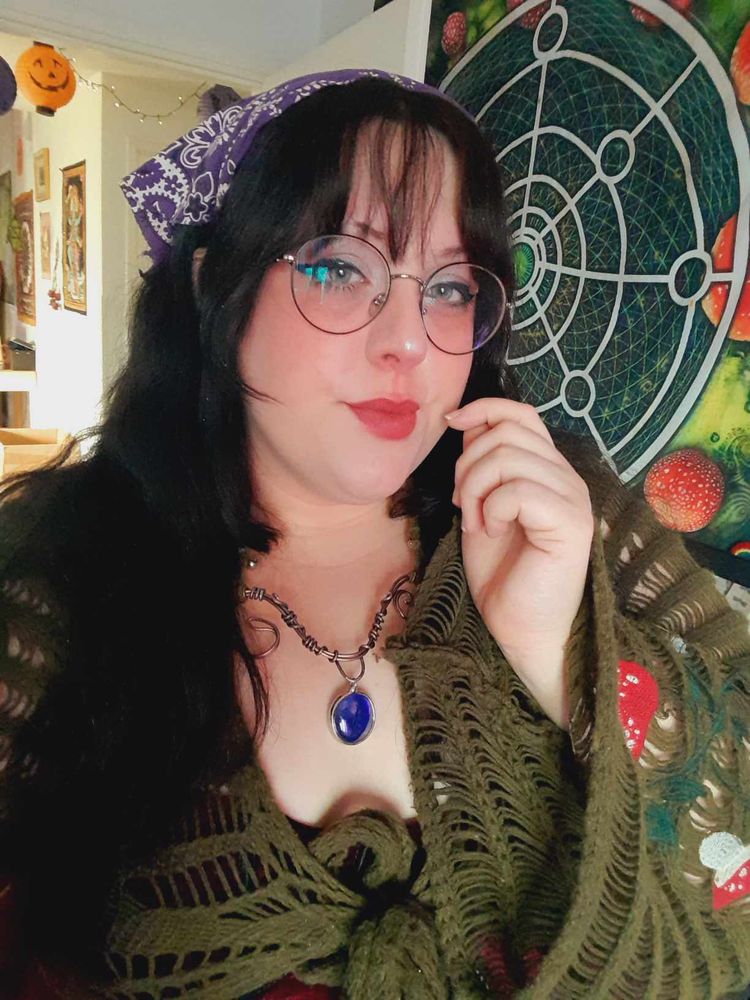 A photo of a girl with long, black hair. She is looking at the camera and her hand is up near her face. She is wearing a purple bandana, round glasses, a knitted dark green sweater with mushroom applique on the sleeves and a swirly metal necklace with a bright blue glass pendant. Behind her is a tapestry hung on a wall with mushrooms in a circle surrounding a large white spiral. 