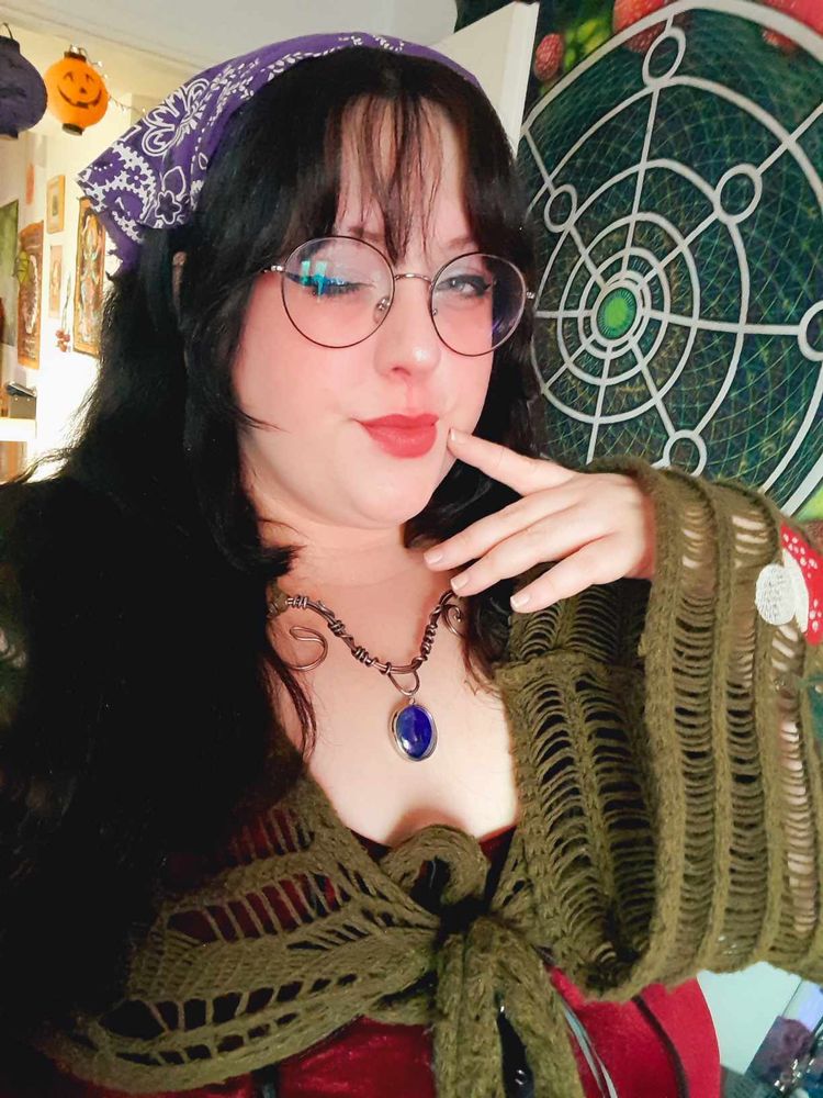 Similar to the other photo in this post, this is a photo of a girl with long, black hair. She is looking at the camera and her hand is up near her face with her index finger resting on her cheek. She is wearing a purple bandana, round glasses, a knitted dark green sweater with mushroom applique on the sleeves and a swirly metal necklace with a bright blue glass pendant. Behind her is a tapestry hung on a wall with mushrooms in a circle surrounding a large white spiral. 