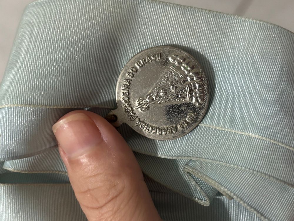 Foto de uma fita azul já amarelada pelo tempo um dedão aparece segurando uma medalha prata de nossa senhora Aparecida. Essa fita é da congregação Mariana.
