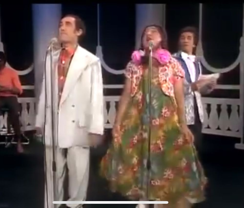 Cena de os Trapalhões em show de calouros Didi veste conjunto social branco e Zacarias vestido florido, peruca com tranças para frente e duas flores rosas uma em cada trança, ao fundo Dede vestindo terno estampado em branco e cinza.
Interpretação de " papai eu quero me casar" 
