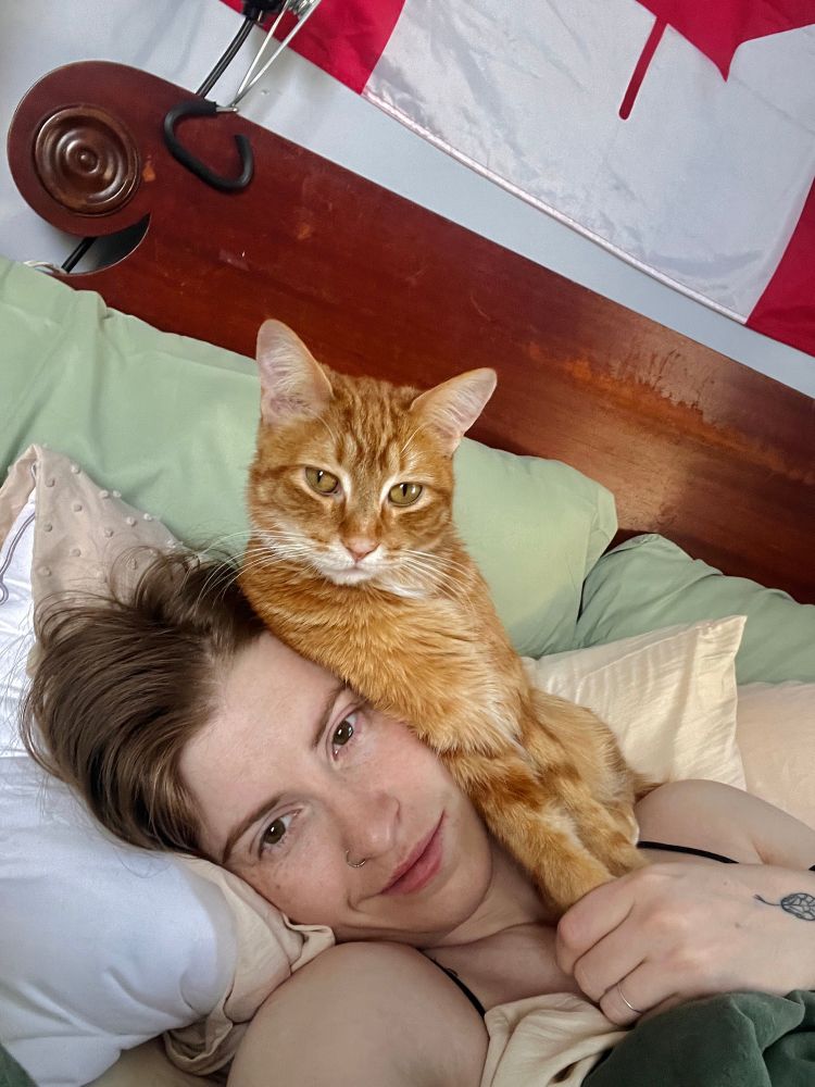 Cooper is laying in bed with her head tilted to the side - and oliver (orange cat) is sleeping on top of her face, with his paws safely held in cooper’s hand. 
