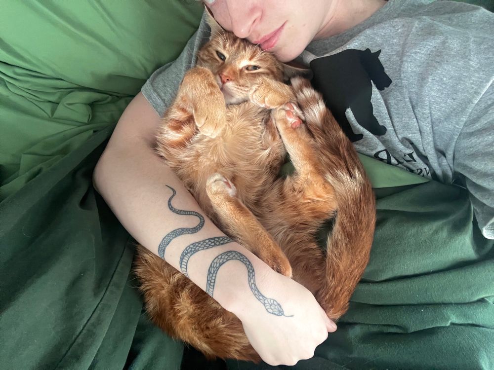 An orange cat on his back, with his paws in the air, tucked into Cooper’s armpit with their arm wrapped around the kitty, holding onto his bum. 