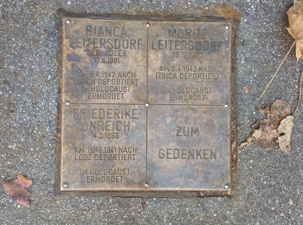 Messingplatte am Boden, mit drei Namen und Daten und der Inschrift "Im Gedenken" aber insgesamt nur schwer lesbar. Ein paar Blätter rundherum. 