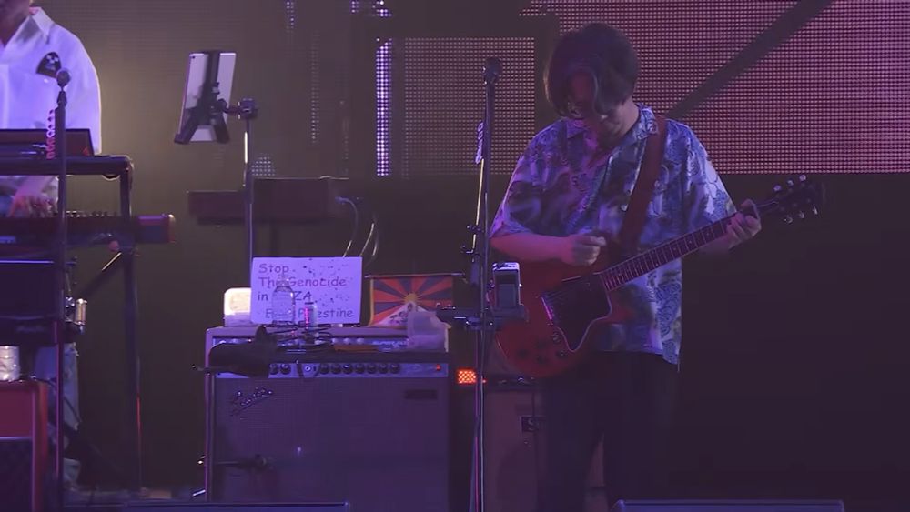 Masafumi Gotoh from Asian Kung Fu Generation with a sign behind him that says

"Stop The Genocide in Gaza. Free Palestine."