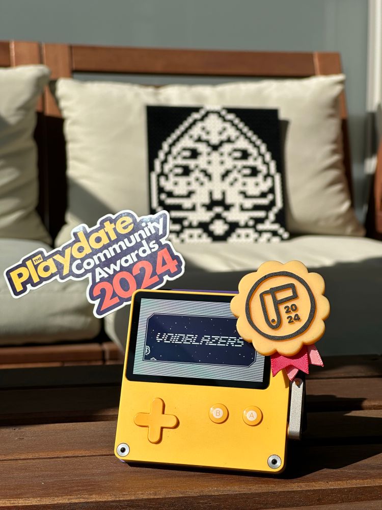 A Playdate showing the title card of “Voidblazers” on its screen. On the left is a sticker that reads “The Playdate Community Awards 2024”. A yellow award rosette sticks to the top right of the device. Cosmeau looms in the background.