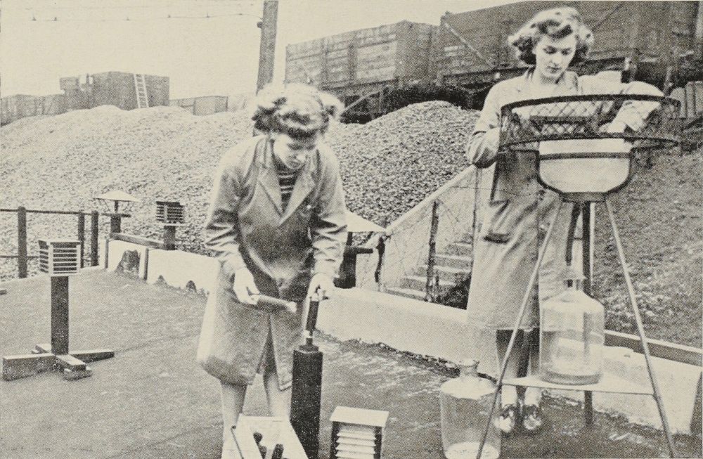 Photographie en noir et blanc. Maintenance par deux femmes vêtues de blouses de laboratoire d'un dispositif de mesure de la pollution atmosphérique placé sur le toit d'un bâtiment non identifié. À gauche, une femme remplace la bougie au peroxyde de plomb pour la mesure du dioxyde de soufre. À droite, une autre femme manipule le pluviomètre employé pour la collecte des particules de charbon.