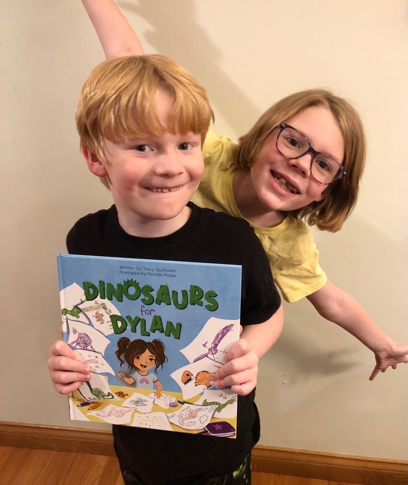 Two cute children, a boy and a girl with pale skin and blond hair, posing with the book Dinosaurs for Dylan