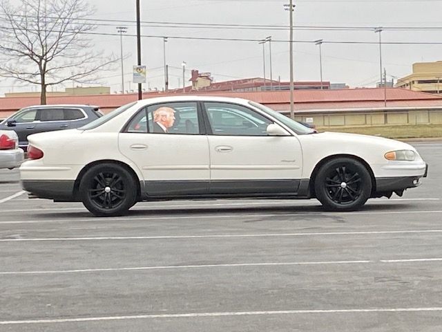 A white car with a sticker of Donald Trumps head on the backseat window as if he is riding in the back seat. 