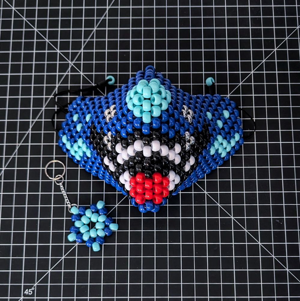 A mask made of blue, black, white, red and silver plastic barrel beads, displayed on a black background with a white grid. The beads are arranged to look like an animal's mouth with its tongue sticking out. Below the mask on the left is a star keychain made with matching blue beads. 