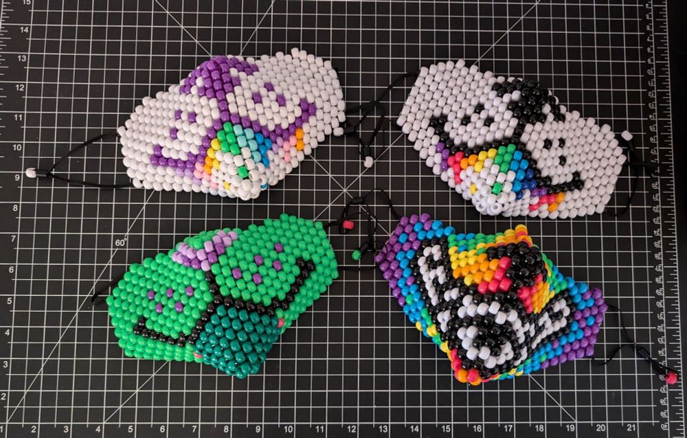 Four beaded masks placed on a black cutting mat with a white grid. The top left mask is made of white, purple and pastel rainbow beads, designed to look like a dog's mouth with rainbow drool dripping from it. The mask on the top right follows the same rainbow drool design, with white, black and neon rainbow beads. The bottom left mask is made with green, purple and black beads to look like a smiling cat's mouth, and the bottom right mask is made with rainbow, black and white beads to look like a grinning beast's mouth. All four masks have black elastic straps attached to their sides, with a matching bead on the end of each strap.