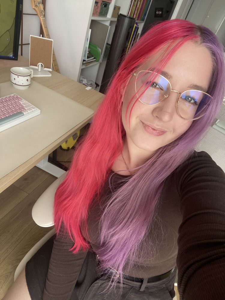 picture of niimsey, smiling, sitting on a chair by the desk. she's wearing dark chocolate brown lshirt and shorts. her hair is straight, with pink and purple split dye. 
