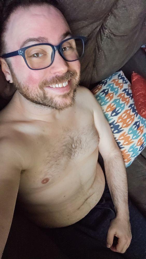 Brandon sitting on his couch wearing just some jogger shorts, smiling into the camera 