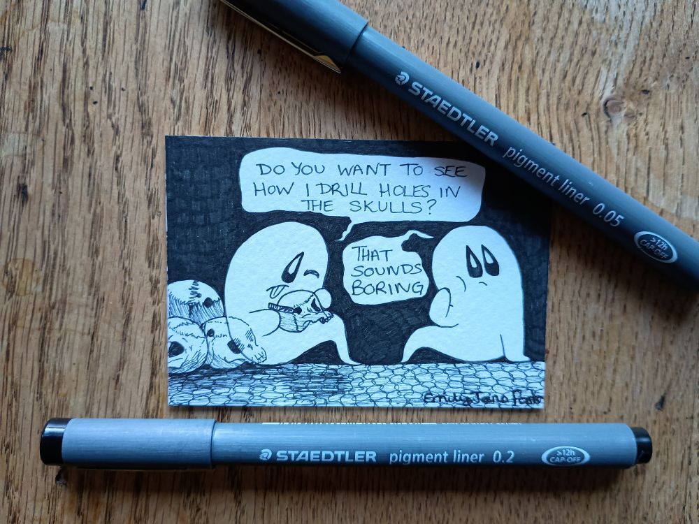 A photograph of an ACEO sized black and white ink illustration on a wooden table with two Staedtler pigment liner pens (a 0.05mm and a 0.2mm). In the illustration, a ghost on the left is sat down leaning back on a pile of skulls. It is hunched over, tongue out in concentration, as it drills a hole in a skull it is holding. The ghost says 