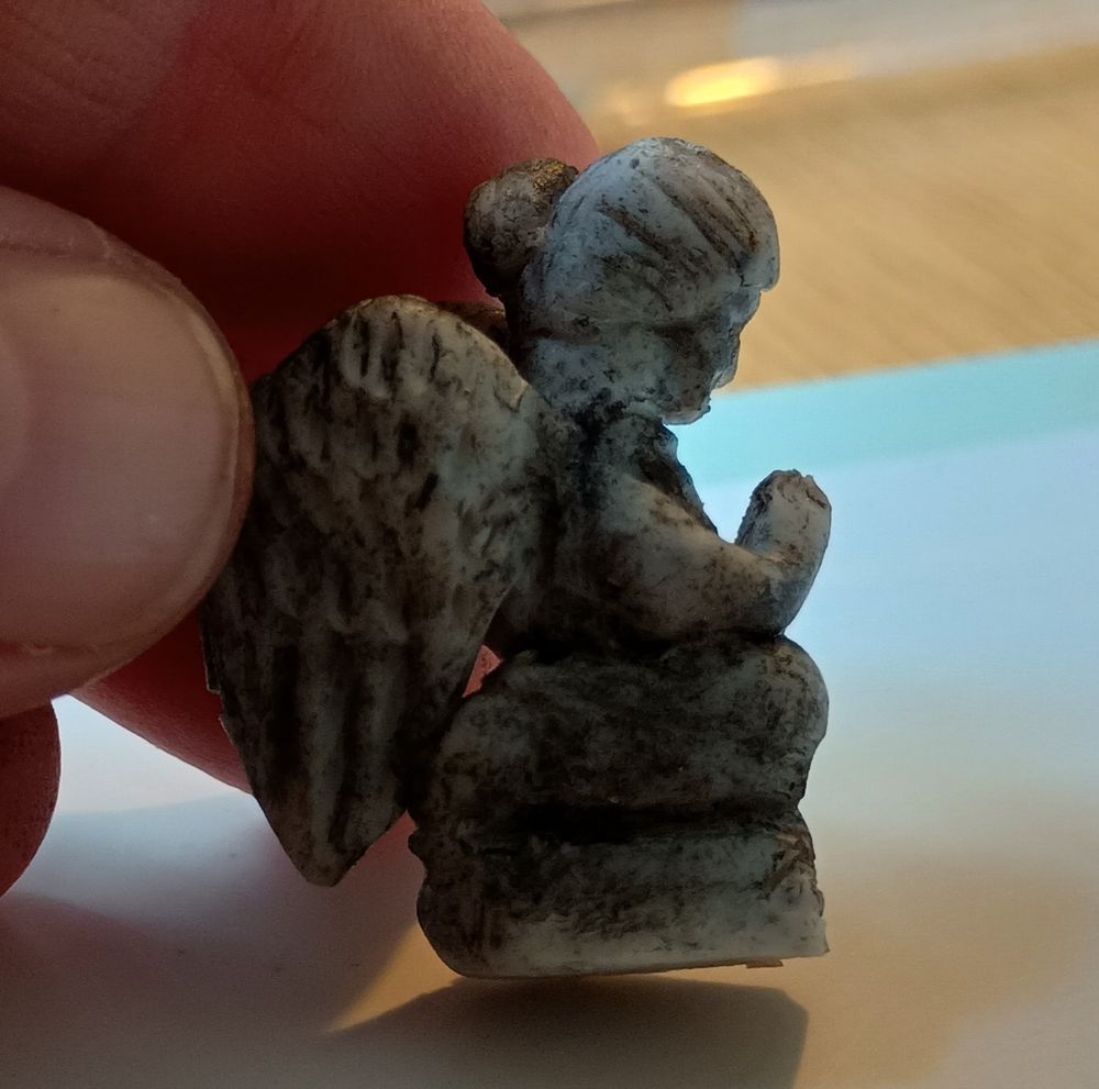 A photograph of a miniature polymer clay angel memorial with acrylic paint dry brushing being held between a thumb and forefinger. It is kneeling in prayer and has hair in a bun.