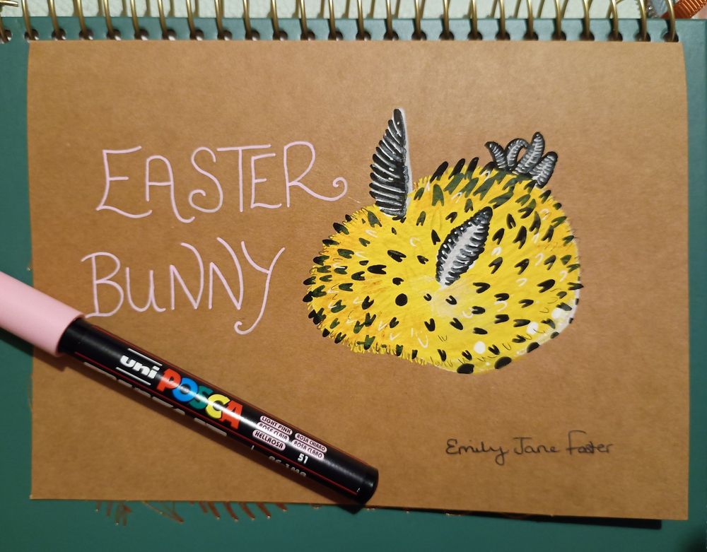 A photograph of a mixed media Posca pen, Staedtler pigment liner and Prismacolour pencil illustration on Kraft card with a Posca pen resting on top of a green and gold embellished planner. The illustration shows a yellow sea bunny, a nudibranch, with the words Easter Bunny next to it. Signed Emily Jane Foster.