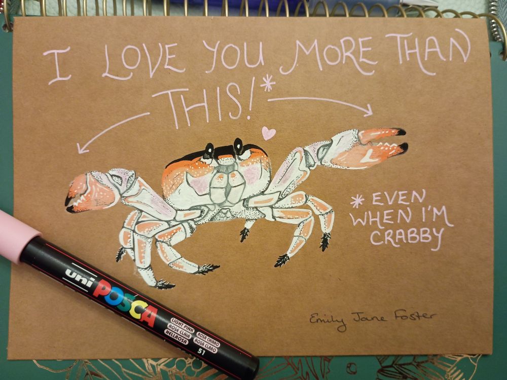 A photograph of an illustration on Kraft card with a Posca paint pen on top of a green and gold embellished spiral bound planner. The illustration shows a blushing crab with claws outstretched and a little heart. The text reads 'I love you more than this!* *Even when I'm crabby'. Art by Emily Jane Foster.