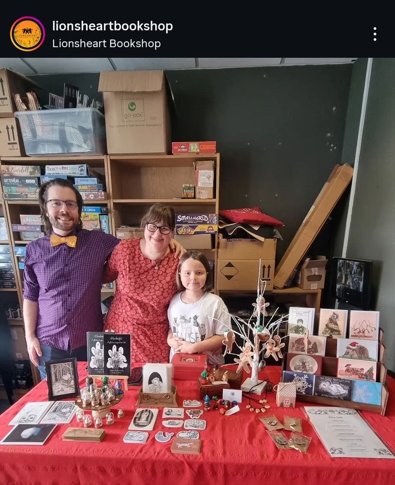 A screenshot of a picture from the Lionsheart Bookshop Instagram page. The photograph shows a family of three standing behind an arts and crafts stall. Behind them are bookshelves with the bookshops boxes. On the stall there are an assortment of wooden carvings, mini polymer clay sculptures in glass terrariums, cards, books, stickers, felt decorations, original art and pin badges.