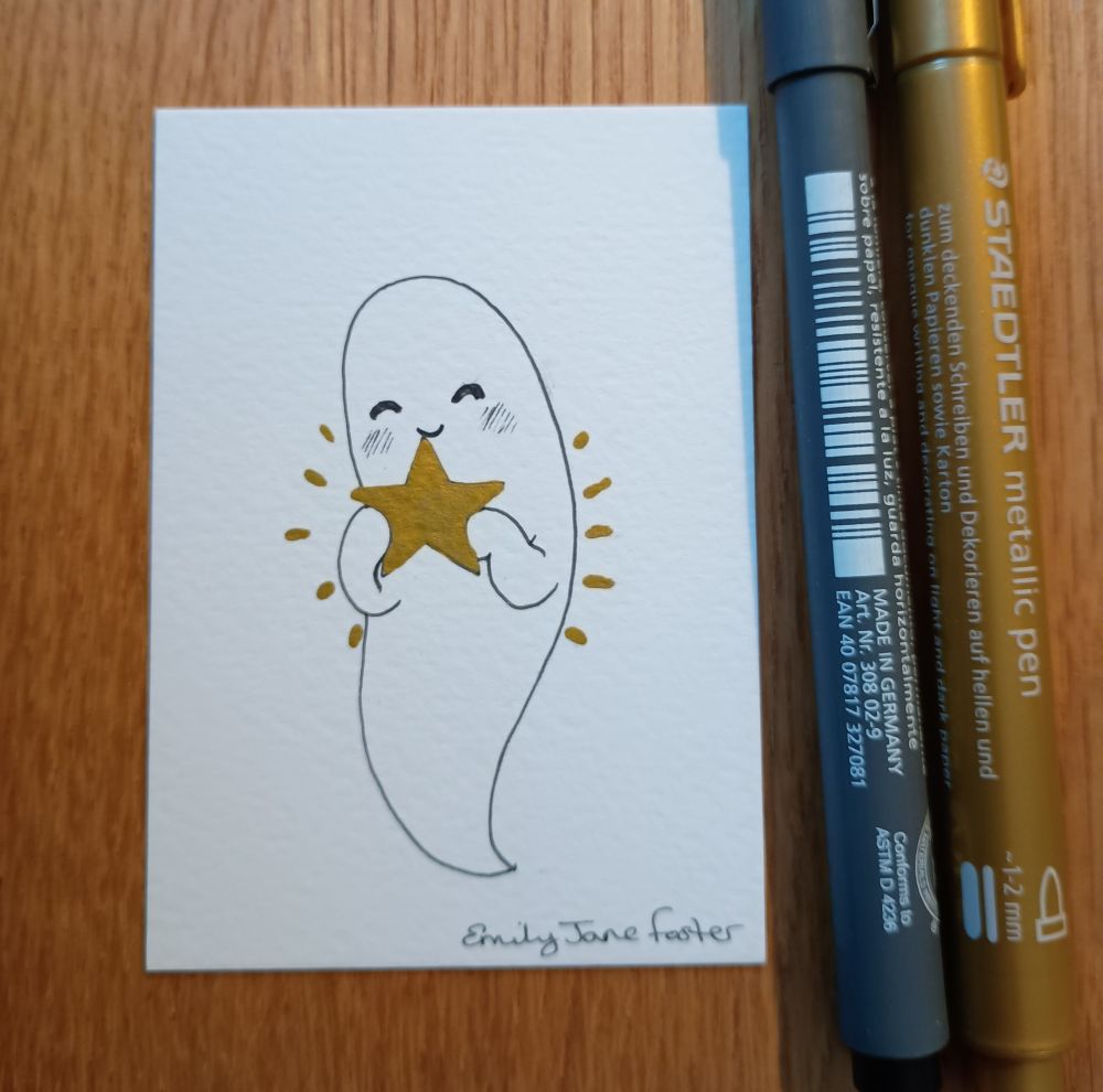 A photograph of an ACEO sized black, white and gold ink illustration on a wooden table with a Staedtler pigment liner pen and a Staedtler metallic pen. A very happy ghost holds out a gold star for you, smiling. Art by Emily Jane Foster.