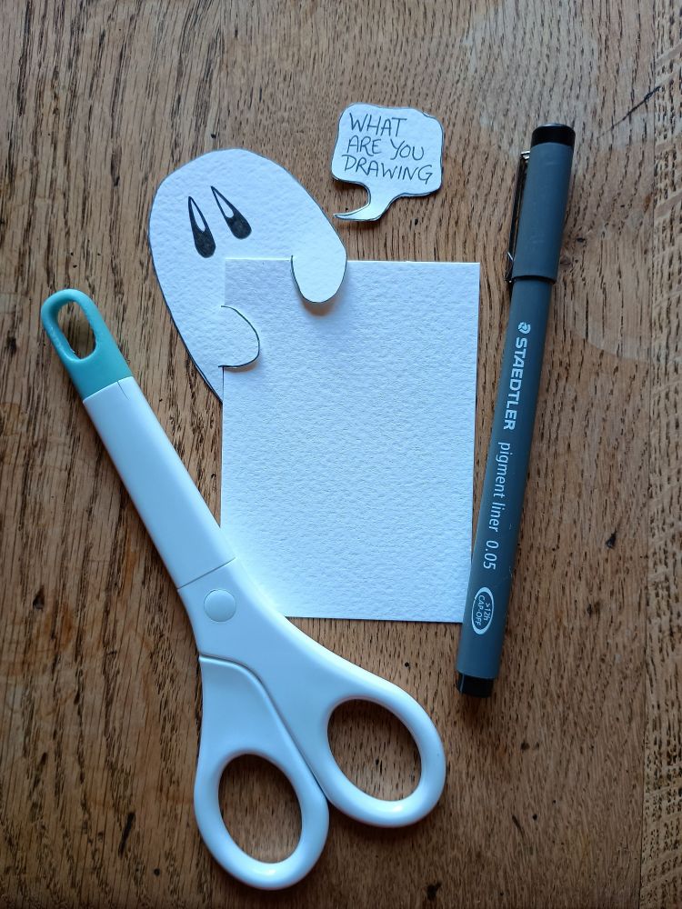 A photograph of a blank ACEO sized piece of watercolour paper. There is a paper craft ghost holding the corner of the paper, with a cut out speech bubble on the table above it saying 